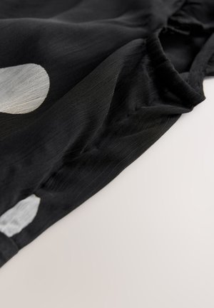 Czarna tkanina z białym wzorem w kształcie kół, charakteryzująca się gładką fakturą i miękkim układem. Detal przedstawia linię dekoltu i obszar ramienia.