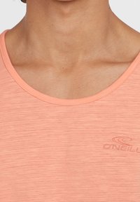 Světle korálové tričko vyrobené z měkké, strukturované látky; má kulatý výstřih a decentní branding. Textura vypadá prodyšně a lehce.