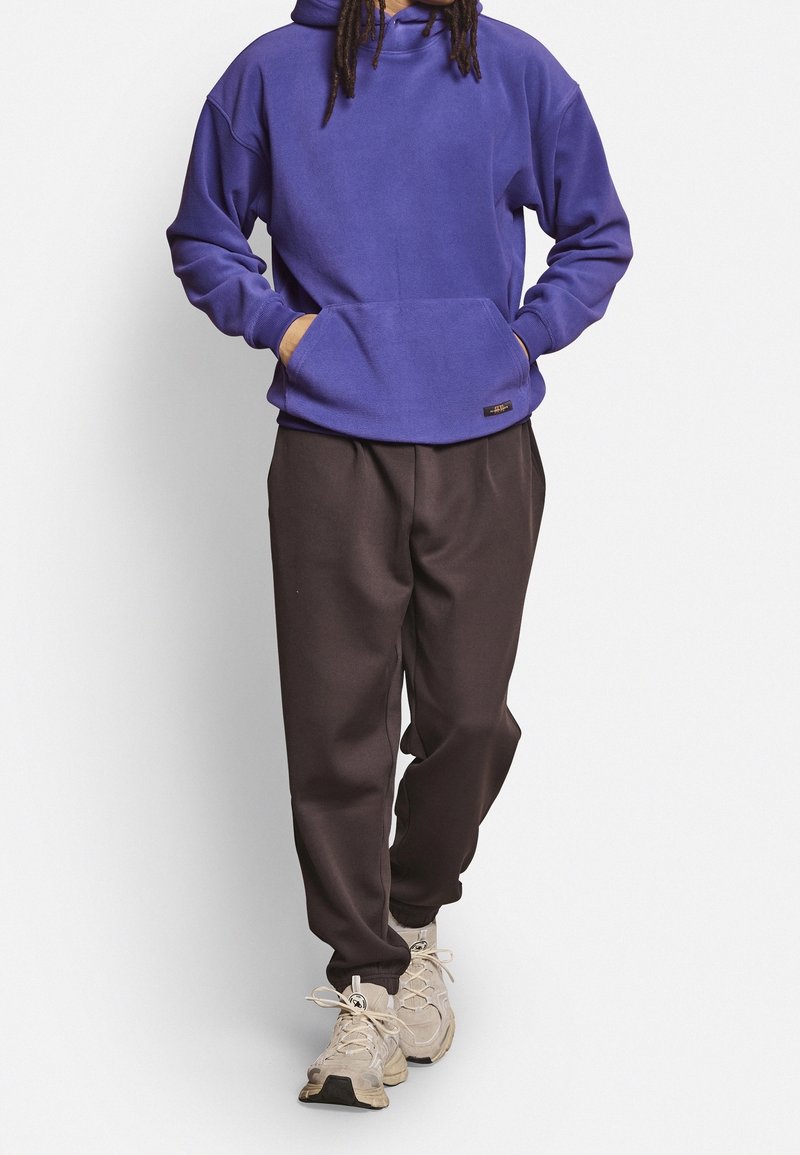 Purple fleece hoodie with a front pocket, paired with dark brown jogger pants and beige sneakers, creating a relaxed outfit.