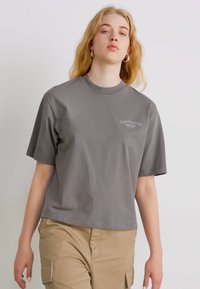 T-shirt grigio a maniche corte in cotone con collo rotondo. Presenta una stampa logo discreta sul petto. Abbinato a pantaloni khaki.