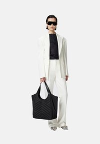 Borsa a tracolla nera trapuntata con motivo a zigzag, dotata di manici neri resistenti. Indossata con un completo bianco sartoriale e una maglia nera.