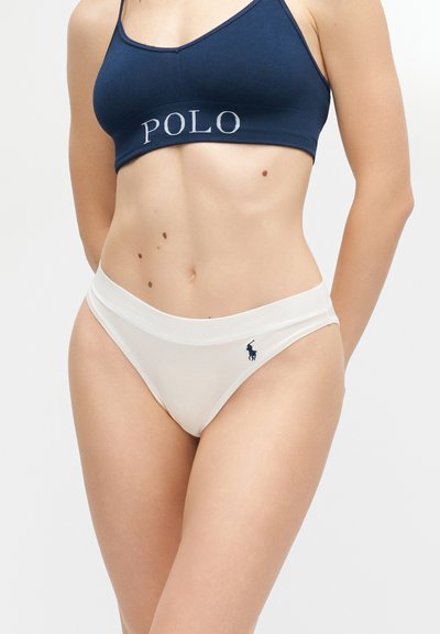 Γυναίκα που φορά ένα σπορ σουτιέν σε ναυτικό μπλε με το κείμενο "POLO" και λευκά σλιπ υψηλής κοπής που φέρουν ένα μικρό λογότυπο παίκτη πόλο στο ισχίο.