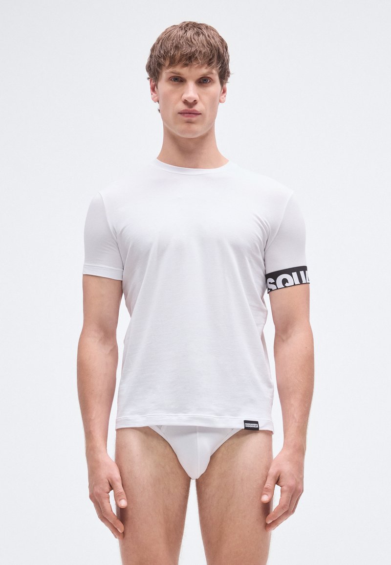 Hvid kortærmet t-shirt lavet af blødt stof, med et sort logo-bånd på venstre arm og et mærke ved kanten. Figursyet design.