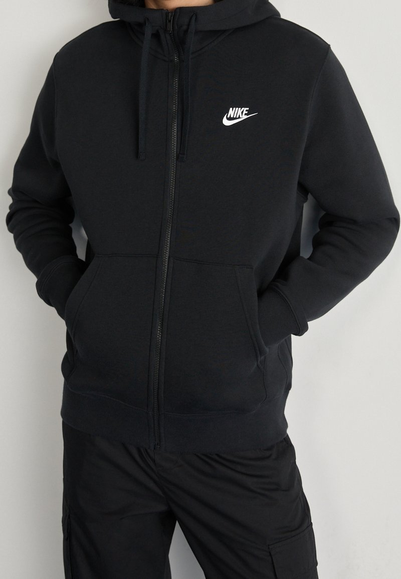 Persona vistiendo una sudadera con cremallera negra de Nike, con las manos en los bolsillos frontales, combinada con pantalones cargo negros, contra un fondo de pared lisa.