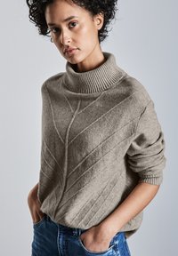 Maglione beige lavorato a maglia con colletto alto a coste, motivi testurizzati diagonali e maniche arrotolate, abbinato a jeans blu.