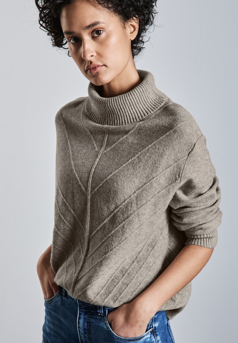 Maglione beige lavorato a maglia con colletto alto a coste, motivi testurizzati diagonali e maniche arrotolate, abbinato a jeans blu.