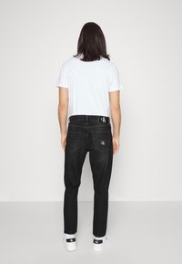 Svarta denimjeans har en slim fit, flera fickor, kontrasterande sömmar och en logotyp på midjebandet. De är kombinerade med en enkel vit t-shirt.
