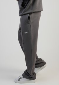 Szürke, puha anyagból készült melegítőnadrág, oldalsó zsebekkel és egy kis fehér logóval a bal combon. Szürke és fehér sportcipővel párosítva.