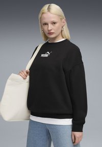 Felpa nera con logo bianco, tessuto morbido, vestibilità rilassata e maniche lunghe. Abbinata a jeans azzurri chiari e una borsa tote color crema.