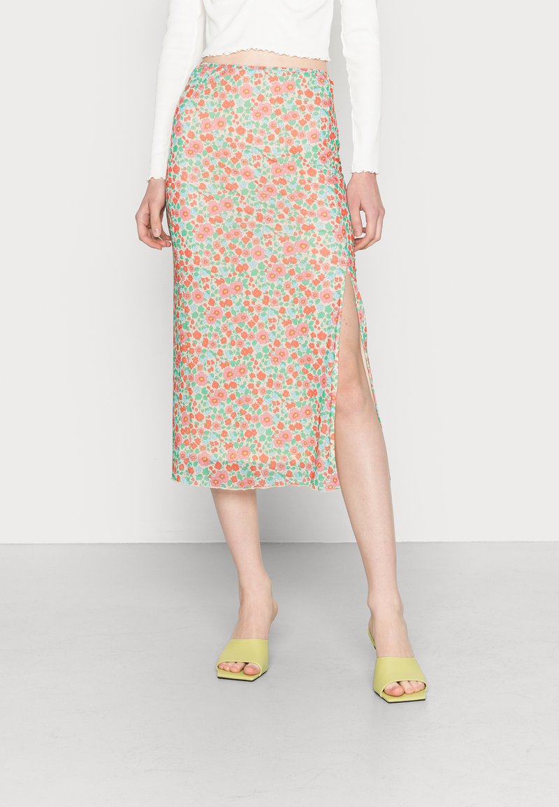 Saia midi floral com fundo verde, flores vermelhas e rosas, fenda lateral, combinada com sandálias de salto bloco verdes.