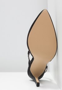 Chaussure noire à talons hauts avec un bout pointu, en suède, talon aiguille fin et semelle beige. La chaussure possède une bride à la cheville.