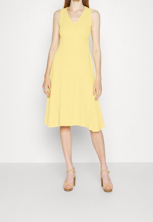 Femme portant une robe jaune sans manches, à encolure en V et longueur au genou, avec des sandales à plateforme beige, debout devant un fond blanc.