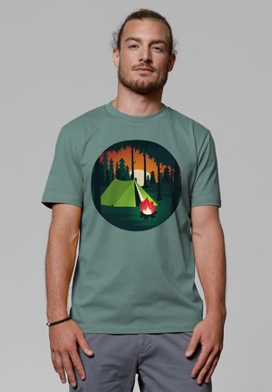 Groene katoenen t-shirt met een ronde afbeelding van een tent en kampvuur, tegen een zonsondergang met bomen op de achtergrond.
