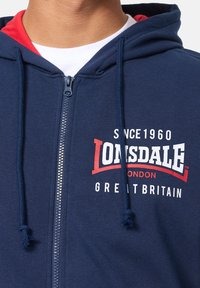 Felpa zip con cappuccio di colore blu navy con accenti rossi, caratterizzata dal logo Lonsdale bianco e rosso. Progettata con cappuccio regolabile e tessuto morbido.