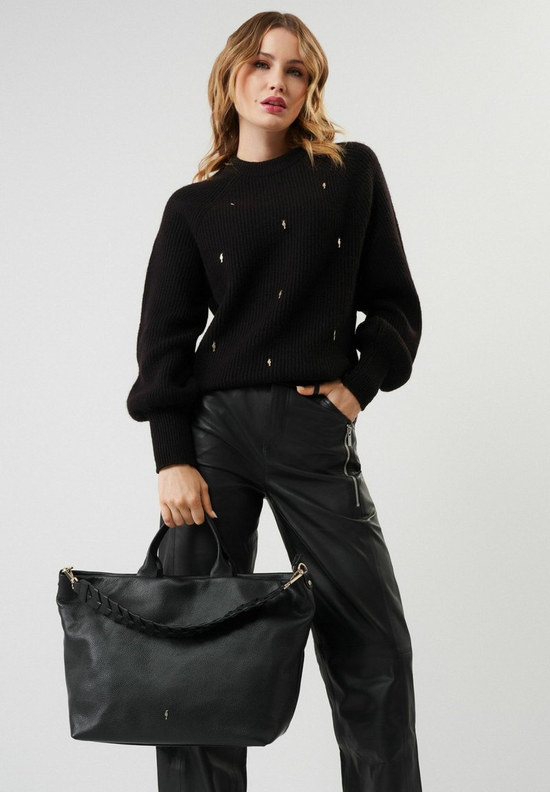 Czarna skórzana torebka o teksturowej powierzchni i plecionym uchwycie, trzymana przez osobę w czarnym swetrze i skórzanych spodniach.