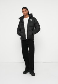 Jeune homme avec des cheveux tressés portant une veste rembourrée noire, un t-shirt blanc, un pantalon noir et des baskets noires, se tenant les mains dans les poches.