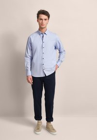 Hellblaue Button-Up-Bluse mit braunen Knöpfen, kombiniert mit dunkelblauen Hosen und beigen Sneakers. Die Bluse hat lange Ärmel und einen klassischen Kragen.