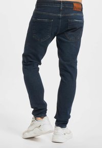 Mann iført mørkeblå, slanke jeans og hvite, chunky joggesko, stående med føttene litt fra hverandre på hvit bakgrunn.