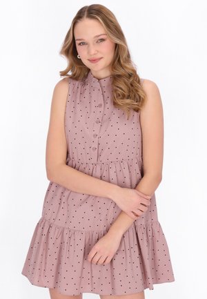 Junge Frau mit welligem Haar, die ein ärmelloses rosa Kleid mit schwarzen Punkten trägt und lächelt, während sie die Arme sanft vor der Brust verschränkt.