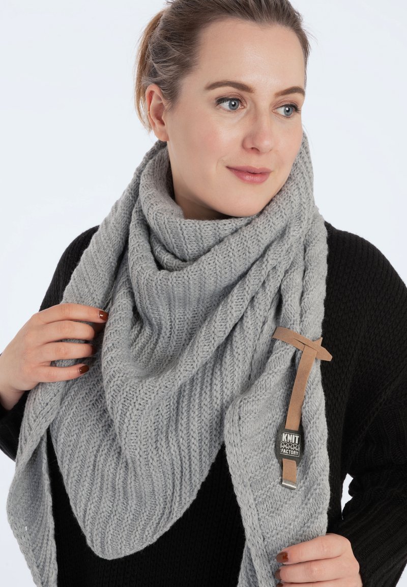 Grijze gebreide sjaal met een gestructureerd ribbelpatroon, gedrapeerd in een driehoekige vorm. Voorzien van een leren label met "Knit Factory" branding.