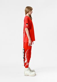 Una camiseta roja de gran tamaño y pantalones a juego presentan gráficos de alas y rayas de contraste. El conjunto se complementa con botas de combate beige y etiquetas decorativas.