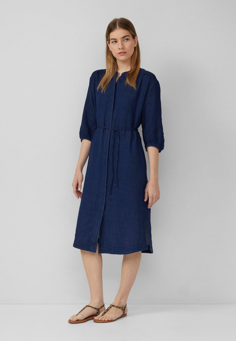Abito blu navy a lunghezza ginocchio con abbottonatura frontale, maniche arricciate, vita con fiocco e spacchi laterali; realizzato in un tessuto leggero.