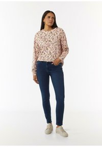 Bloemige top met lange mouwen op een roze achtergrond, donkerblauwe skinny jeans en lichtgekleurde sneakers. Geplaatst tegen een effen achtergrond.