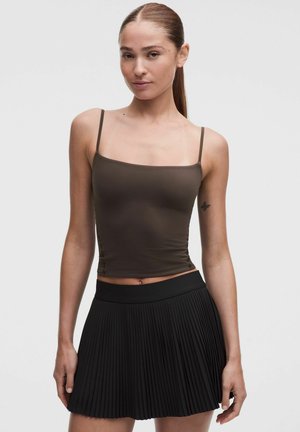Joven mujer con camiseta sin mangas marrón y falda negra plisada de pie contra un fondo claro y liso.