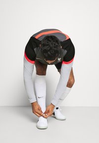 Ropa deportiva que presenta una camiseta de manga larga gris con acentos en rojo y negro, combinada con calcetines y zapatos blancos, enfocado en atarse los zapatos.