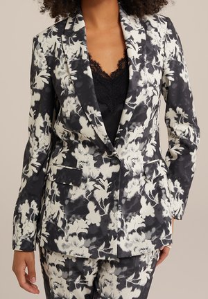 Vrouw draagt een zwart-wit bloemenpak met een colbert met één knoop en bijpassende broek over een zwart topje met kanten rand.