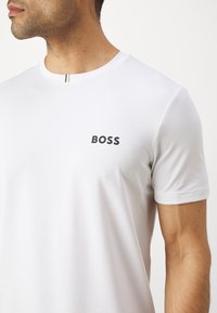 Valge lühikeste varrukatega T-särk, mis on valmistatud siledast kangast; rinnal on peen BOSS logo ja kaeluses õhuke aktsentjoon.