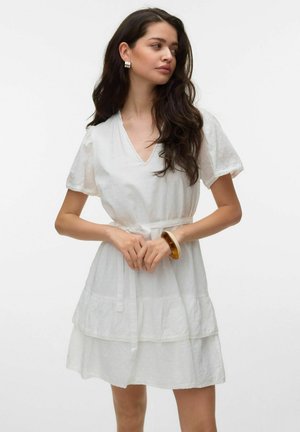 Jeune femme aux longs cheveux foncés portant une courte robe blanche à manches bouffantes et un décolleté en V, accessoirisée de boucles d'oreilles créoles et d'un bracelet.