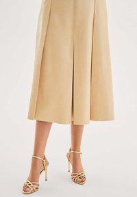 Beige midi-hame, jossa on jämäkkä siluetti ja etuhalkio. Yhdistetty kultaisiin korkokenkiin, jotka korostavat tyylikästä muotoilua.