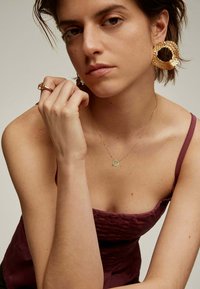 Pendientes de aro texturizados en oro, un delicado collar de cadena dorada con un colgante circular verde y una blusa ajustada color burdeos. Textura de piel suave visible.