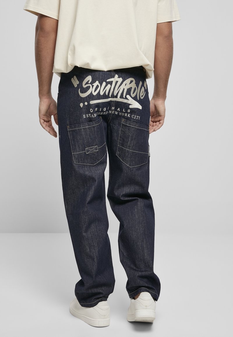 Pantalon en denim bleu foncé avec texte imprimé blanc "SouthPole Originals" au dos, coupe décontractée, design à cinq poches.