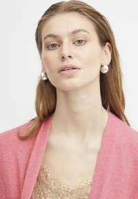 Cardigan rosa sopra un top in pizzo beige, con orecchini in argento di grande impatto. Il modello ha i capelli lisci e un trucco neutro.