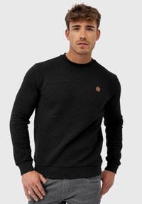 Sudadera negra texturizada con cuello y puños acanalados. Presenta un pequeño parche de logo marrón en el pecho. Modelo vistiendo jeans grises.