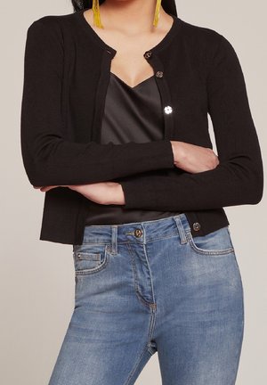 Cardigan noir à côtes avec un col rond, doté de fermetures à boutons brillants, associé à un jean en denim bleu. Texture de tissu lisse et design ajusté.