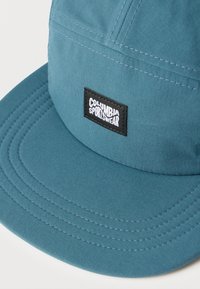 Τσίλινος σκούφος Columbia Sportswear σε τιρκουάζ χρώμα με ραμμένα πάνελ και μαύρη ορθογώνια λογότυπη ετικέτα με λευκό κείμενο στο κέντρο μπροστά.