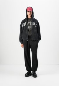Person wearing black hoodie with "NIGHTMARE" text, skull graphic T-shirt, black pants, chunky black boots, and a red baseball cap with hood up.