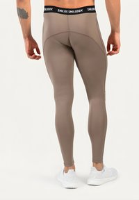Mann trägt taupefarbene Sportleggings und weiße Sneakers, steht und ist von hinten vor einem schlichten hellen Hintergrund zu sehen.