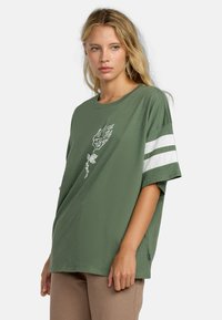 Olijfgroene oversized T-shirt, korte mouwen, met witte strepen op de mouwen en een bloemenafdruk op de voorkant. Katoenen materiaal.