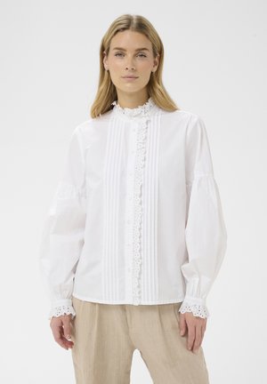 Vrouw met steil blond haar, gekleed in een wit blouse met lange mouwen en kanten details, en beige broek, tegen een effen achtergrond.