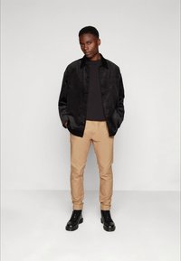Casaco de camurça preto com fecho de botões, combinado com uma camisa preta e calças chino bejes. Finalizado com botas de cano médio pretas. Design simples, sem padrões.