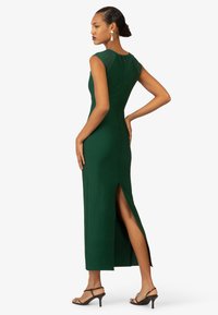 Grünes, figurbetontes Kleid mit hohem Schlitz, kurzen Ärmeln und strukturierten Oberteil-Details. Getragen mit schwarzen Riemchenschuhen, die eine schlanke Silhouette betonen.