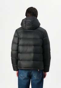 Zwarte pufferjack met een capuchon, met een gewatteerd ontwerp, een gladde textuur en een getailleerde silhouette. Draagt over blauwe spijkerbroeken.