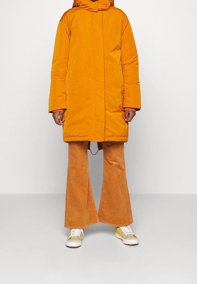 Personne portant un manteau matelassé jaune moutarde, un pantalon évasé en velours côtelé orange et des baskets beiges à lacets blancs, debout devant un fond uni.