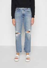 Jeans de mezclilla de color azul claro con detalles de rodillas desgastadas, corte de pierna recta, cintura de subida media y dobladillo deshilachado, combinados con sandalias blancas tipo slide.