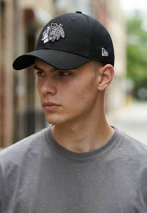 Junger Mann mit schwarzer Netz-Baseballkappe mit weißem Indianer-Emblem, der zur Seite schaut, trägt ein graues T-Shirt mit Rundhalsausschnitt.