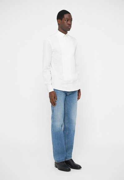 Λευκό πουκάμισο μακρυμάνικο με λεπτομέρεια σε πατέντα, συνδυασμένο με ανοιχτόχρωμα τζιν και μαύρες δερμάτινες παπ�ούτσια, παρουσιασμένο σε λευκό φόντο.